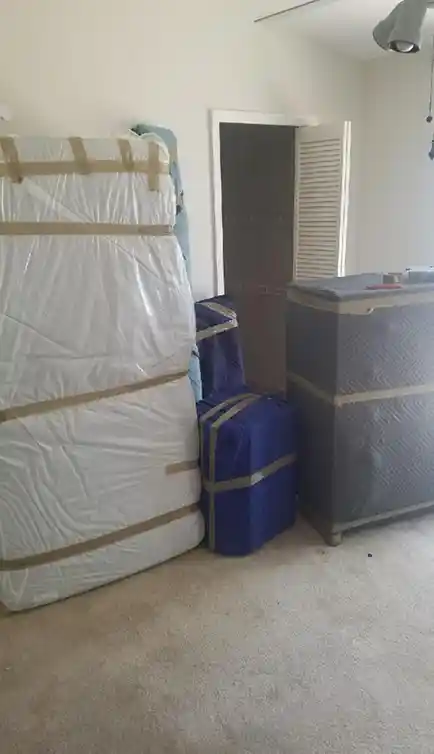 Several large household items, including mattresses and furniture, are wrapped and taped with protective coverings, stacked against a wall in a carpeted room. An open door is visible in the background.