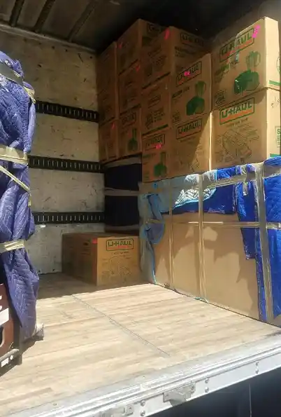 Several packed and sealed U-Haul moving boxes stacked neatly inside a moving truck, with furniture wrapped in blue blankets and straps securing the items in place.